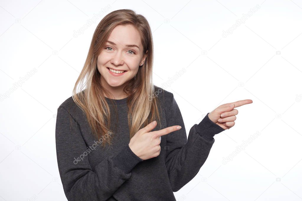 Studio isolated portrait of positive blonde woman wears grey casual sweater, has pleasant broad smile, indicates with hands and fingers at copy space for your advertising content on white background.
