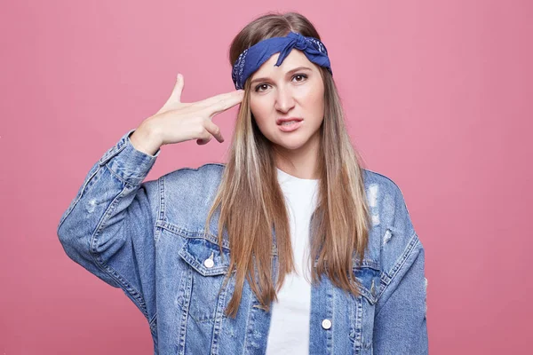 Çekici sıkılmış genç bayan portresi Tapınağı'nda parmak tutar, intihar süsü, trendy var. saç modeli, şık denim içinde giyinmiş pozlar pembe arka plan boş kopya alanı ile karşı.