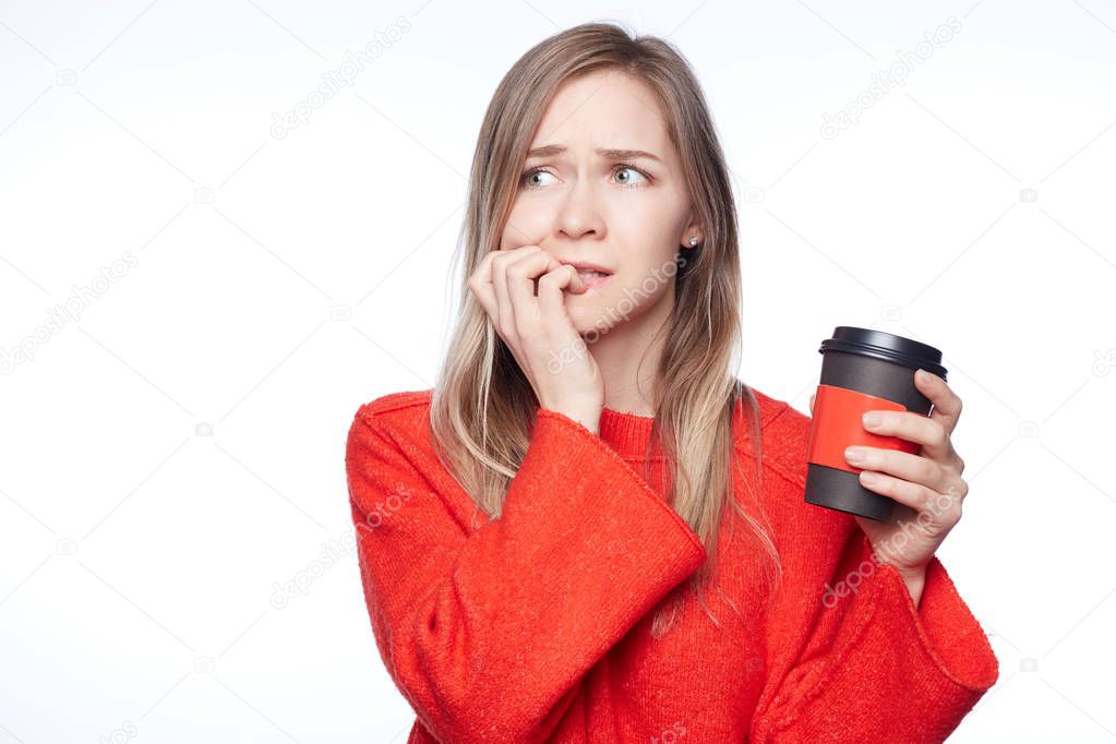 Anxious nervous blonde woman bites finger nails with anxiety, feels worried , wears casual red sweater, poses against white background. Lovely European female with coffee cup in hand has trouble.