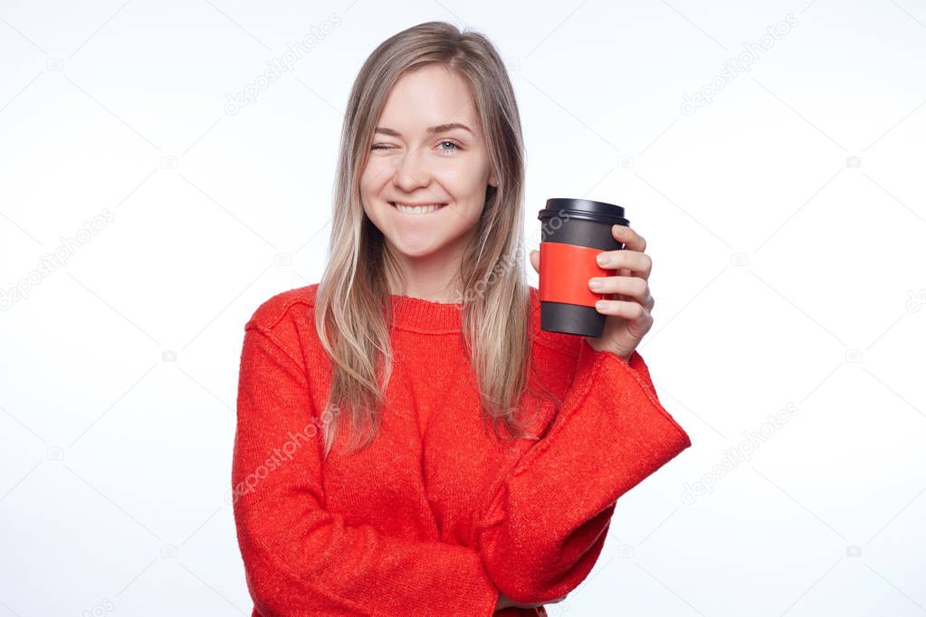 Portrait of beautiful woman with long hair winking, having good mood smiling showing white perfect teeth. Blond young female blinking her eye showing her sympathy or positiveness,  holding coffee cup.