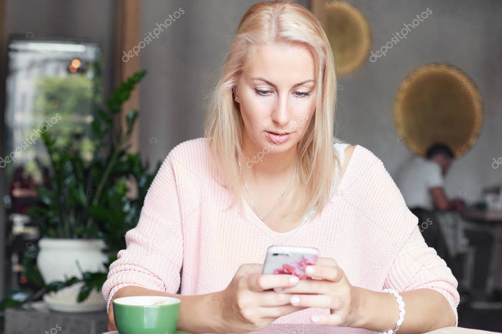 Serious blond European female reads income message with serious expression, checks email online on smart phone, bloggs via modern cellular, spends free time at outdoor cafe. Technology and lifestyle.