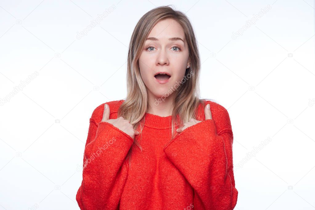 Headshot of young woman in red holding hands on breast, verbally defending herself, having perplexed and puzzled faceexpression, saying: Who me? Human emotions, feelings, reaction and attitude concept