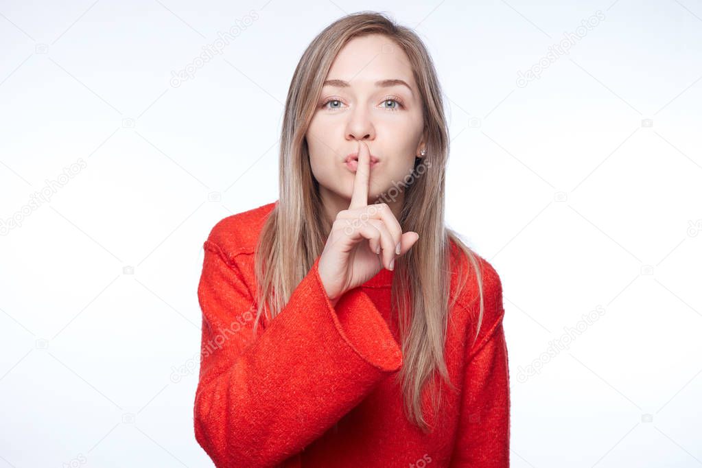 Please, remain silent! Attractive female keeps index finger on lips, makes silence gesture, says shh, asks to be quiet, stares at camera, has long straight blond hair, dressed in fashionable sweater.