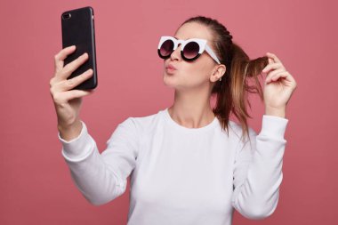 Cep telefonu kullanarak, bir öpücük gönderme otoportre yapma beyaz uzun kollu t gömlek ve şık gözlükler, giyen güzel kızıl saçlı Bayan portresi. Genç erkek pembe duvara fotoğraf çekmek. 
