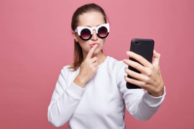 Amerikan çekici kadın yüz buruşturma yapar, selfie için pembe stüdyo duvar arka plan üzerinde pozlar, iş veya eğitim, son moda gözlük giyen ve sosyal ağ için hikayeler yapmak sonra boş zaman var..