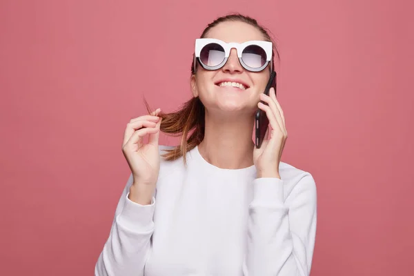 Indoor shot of inspired female student laughs happily during mobile conversation with close friend, enjoys leisure, sits at kitchen, uses modern electronic gadgets. Graphic designer has break