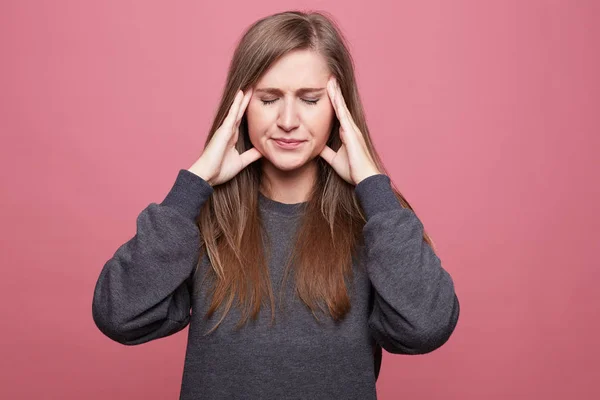 Portrait of European woman has headache, keeps hands on temple, closes eyes, dressed casually, isolated on pink. Desperate frustrated female has troubles, concentrates on solution.Face expression