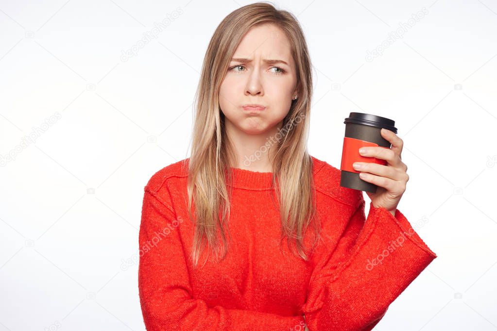 Indoor shot of beautiful European female in red style clothes with coffee cup, blows cheeks, stares with popped eyes away from camera, amazed by shocking news, isolated on white. Facial expressions.