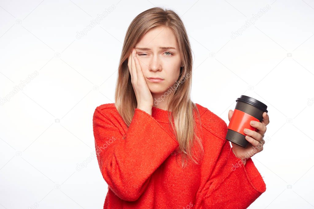Sleepy attractive female student worked all day at diploma paper, poses indoors with cup of coffee, wants to have rest, being tired and exhausted. Exhausted woman in red sweater is tired from work.