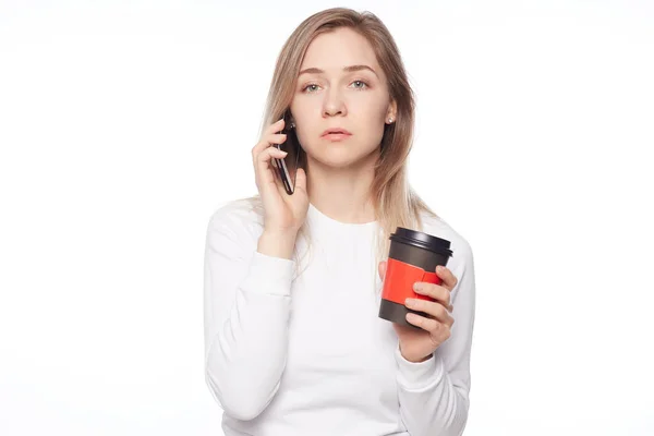 Hate mornings. Indoor shot of tired attractive European female, holding cup of coffee, looking at camera with tired face expression, feeling sleepy and exhausted, standing in office, talking by phone.