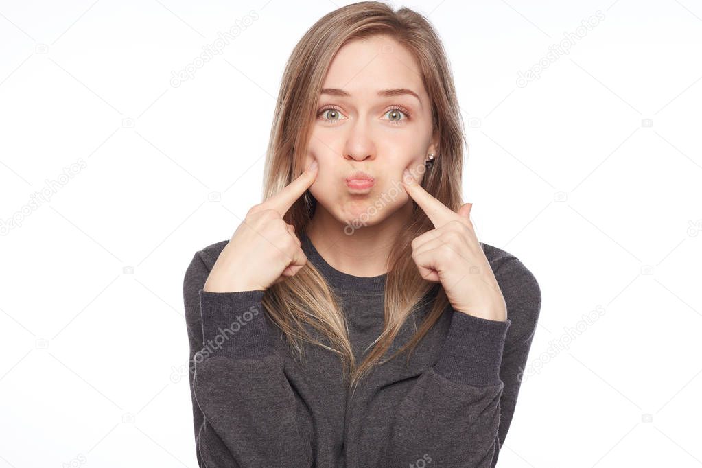 Indoor shot of Caucasian pretty blond woman making grimace blowing cheeks, having pleased face expression. Beautiful young woman with clean tender skin dressed in casual clothes mocking at camera.