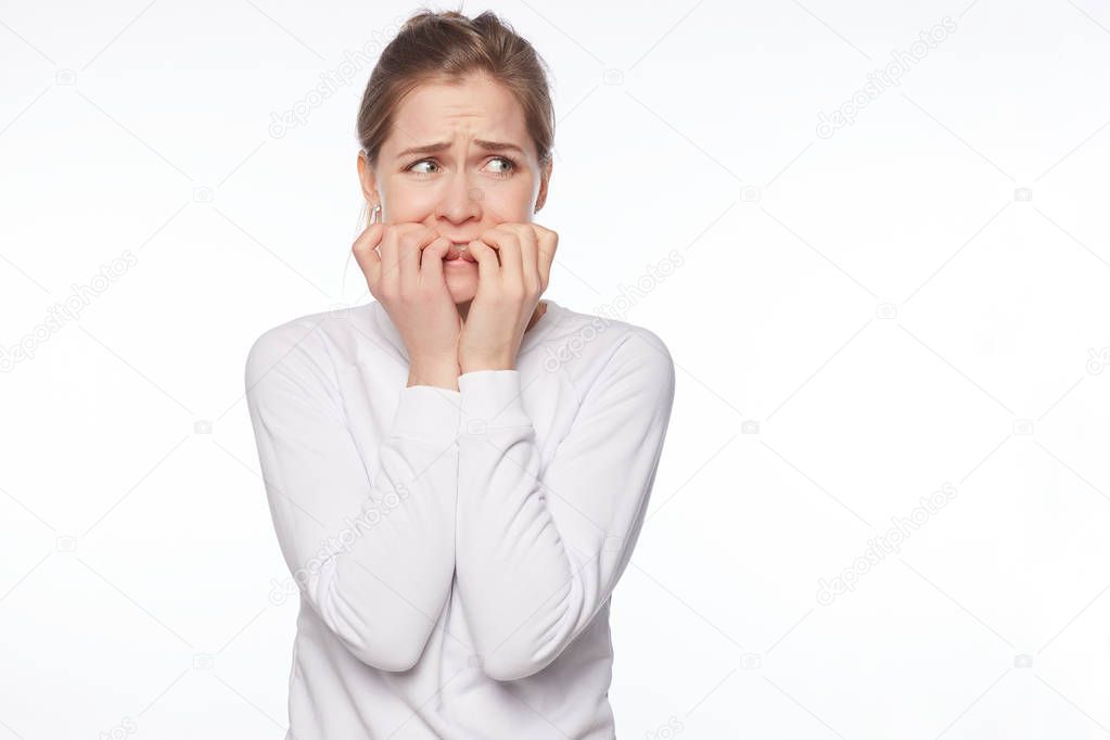 Portrait of scared cute insecure woman with blond hair and grey eyes, biting fingernails and staring around with fear, standing worried and nervous, feeling scared while posing over white studio wall.