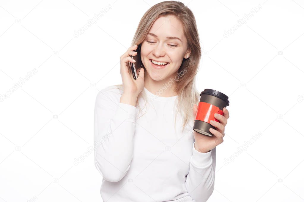 Woman enjoying spending time laughing over new memes. Portrait of beautiful creative blond haired lady in trendy t shirt, drinking coffee and listening funny stories from friend by phone on white wall