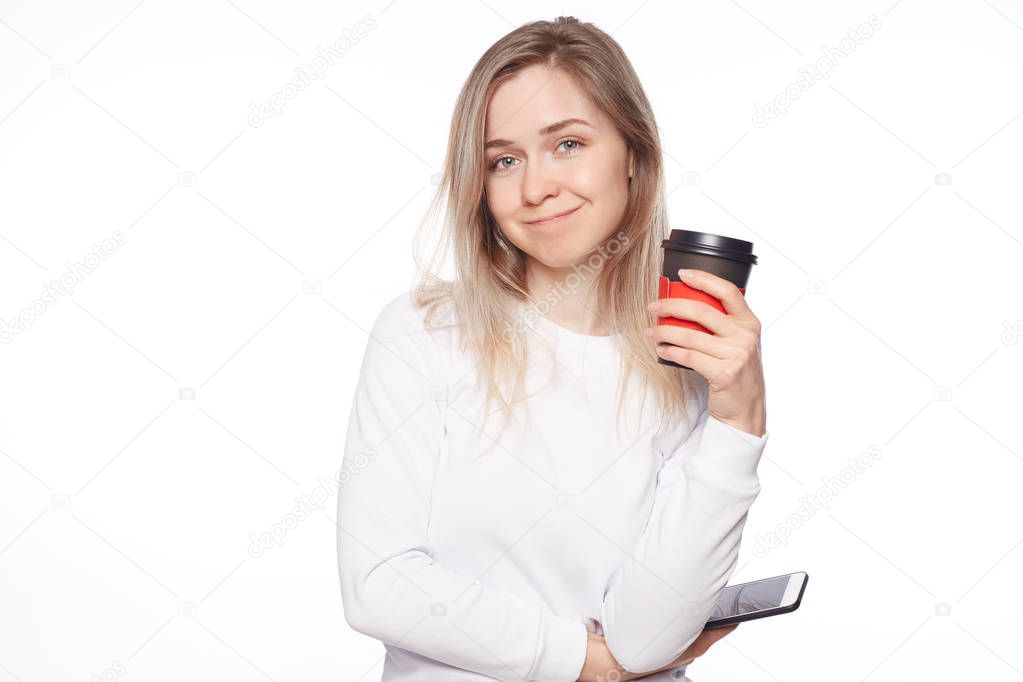 Great day to be productive. Good-looking feminine female in shirt and long hairstyle, looking at camerat with sensual smile, holding cup of coffee, having break during lunch at work over white wall.
