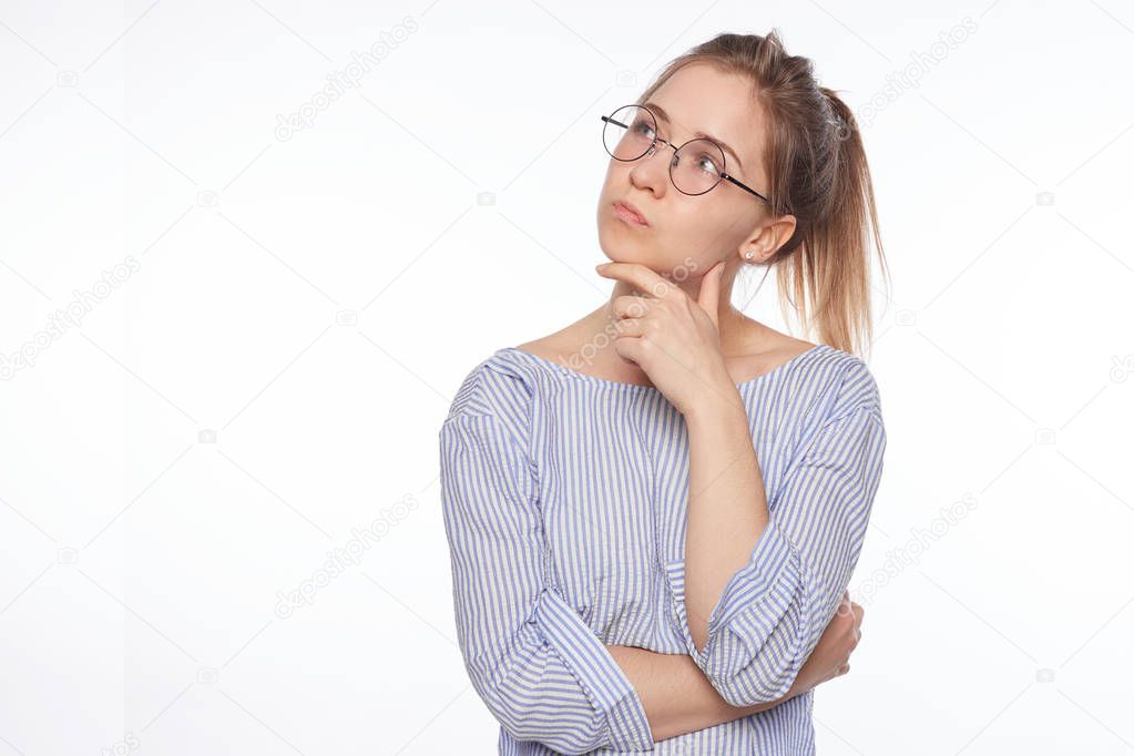 Portrait of beautiful female model with pony tail, wears big spectacles and casual white and blue stripped blouse, keeps hand under chin as thinks about something, poses against studio with copy space