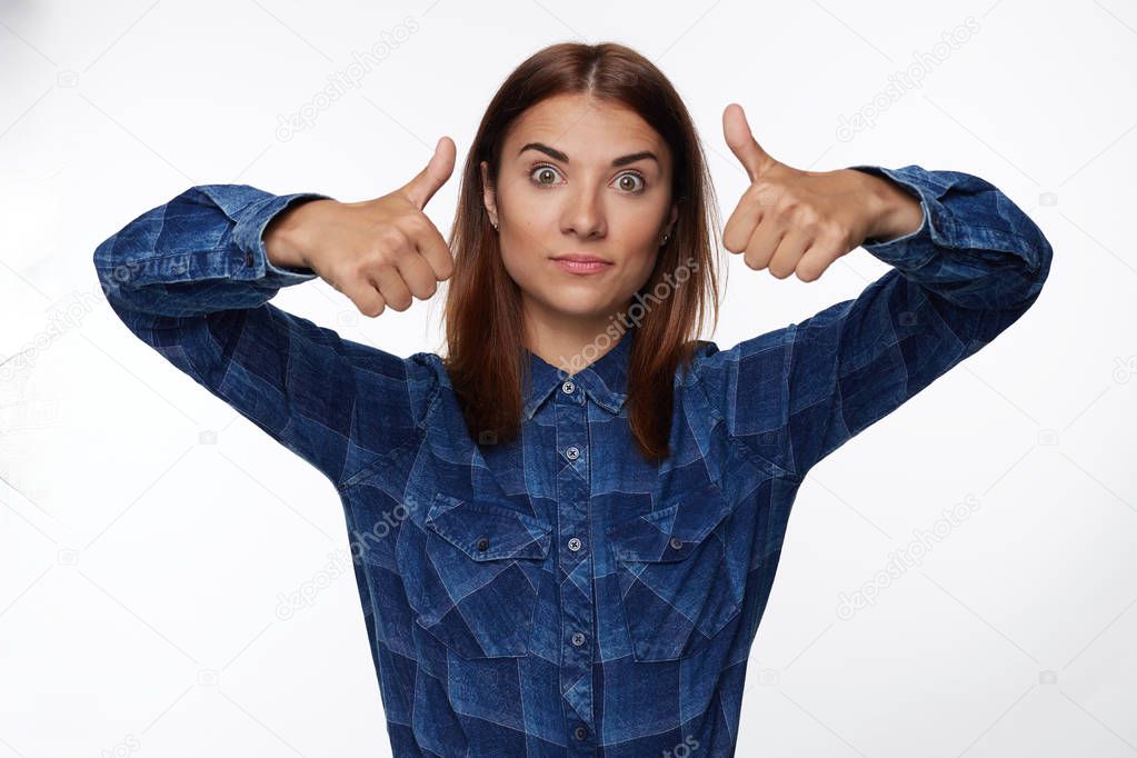 Close-up portrait of young attractive surprised dark-haired woman with bugged brown eyes, wearing casual plaid shirt and looking at camera, showing thumbs up. Isolated on white studio wall background.