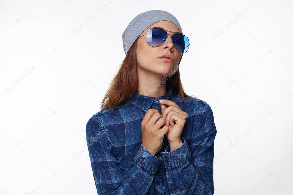 Portrait of stylish attractive young mixed raced female model with straight hair in grey hat and checkered shirt, keeping nose up, looking in camera through blue sunglasses with cool, calm expression.