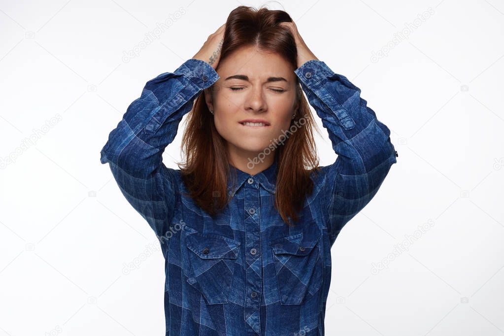 Candid shot of attractive American female closing eyes with worried expression, bites lip, feels anxious and nervous as being scared of something or sees phobia, poses against white studio wall. 