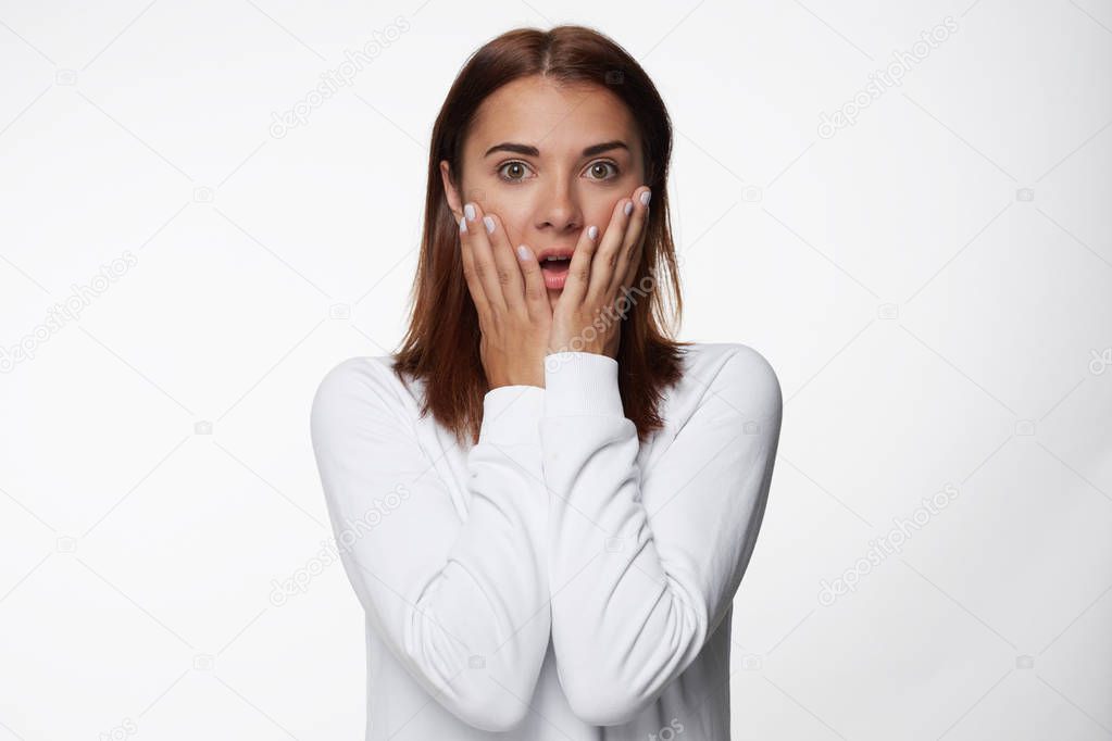 Close up shot of attractive young female with stunned expression, touches cheeks with both palms, stares at camera, being scared of phobia, isolated over white background. Fear and people concept.