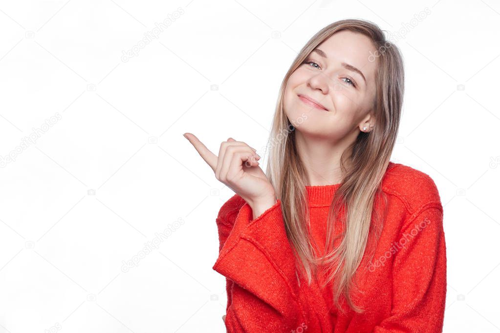 Satisfied glad young blond woman with charming smile points at upper left corner, shows blank space for promotional content, advertises something, wears stylish red loose sweater, isolated on white.