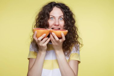 Güzel beyaz kadın ağız ve mavi gözlerini aç yüz ifadesi ile açarak greyfurt, ısırmaya gidiyor. Mutlu genç kıvırcık saçlı kadın yaz giysileri, sarı stüdyo arka plan üzerinde poz giyer