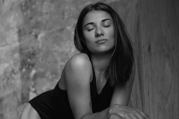 Black and white horizontal shot of Caucasian woman with shot brunette hair, closing eyed, posing indoors, put hands on wooden table. Fashionable shot of young sensual female model.