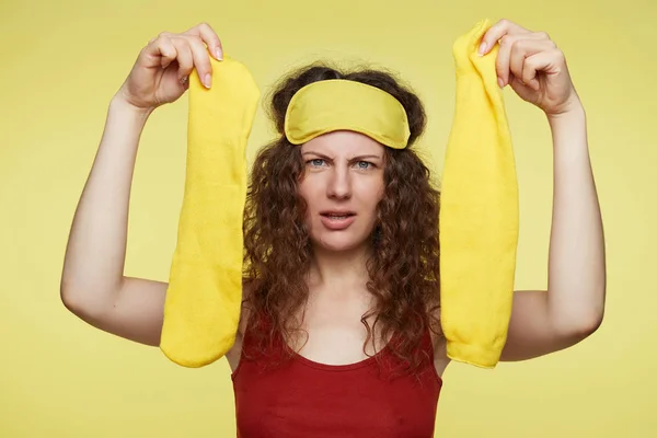 Cute European female passenger opens amenity kits on long-haul international flight frowning forehead, has wondered face expression. Pretty woman wth sleeping yellow mask on head and socks in hand.