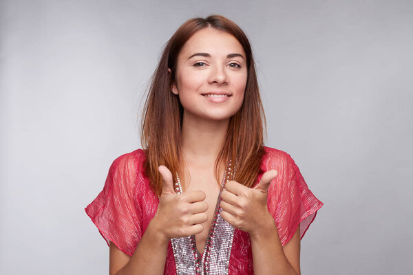 Portrait of young Caucasian female employee or customer with charming smile, looking at camera with happy expression, showing thumbs-up with both hands, achieving career goals. Body language concept.