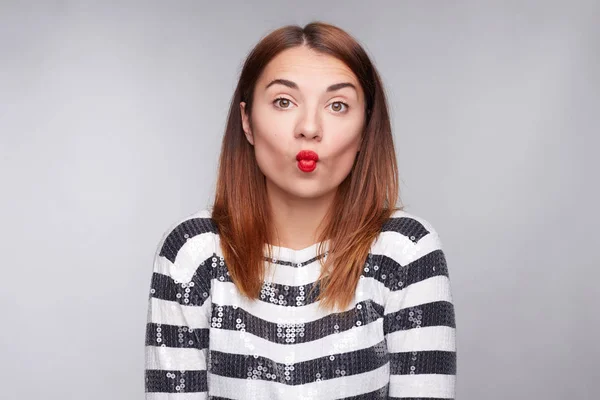 Portrait of young attractive Caucasian hipster woman posing on gray studio wall showing red lips. Student female with short  hairstyle having surprised face expression, staring at camera with grimace.