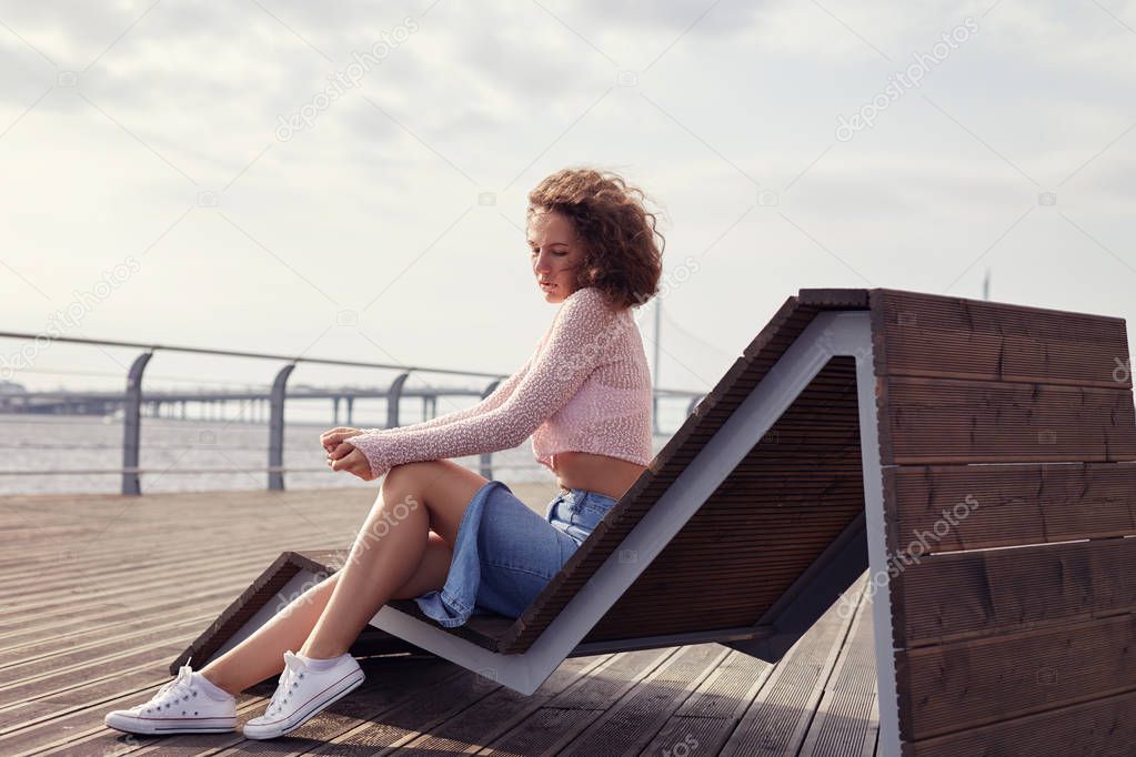 Attractive Caucasian young lady with bushy hairstyle , wearing fashionable casual denim skirt, sitting on wooden sun bed in city park on embankment, looking down, having hurt look. Human emotions.