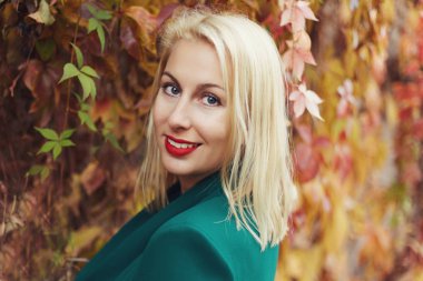 Otantik Genç beyaz bir derin mavi gözlü çok güzel sarışın kadın görüntüsünü kapatmak, kırmızı dudaklar, üzerinde yeşil ceket, kameraya, gülümseyen sonbahar yaprakları renkli doğal duvardan poz yapmak.