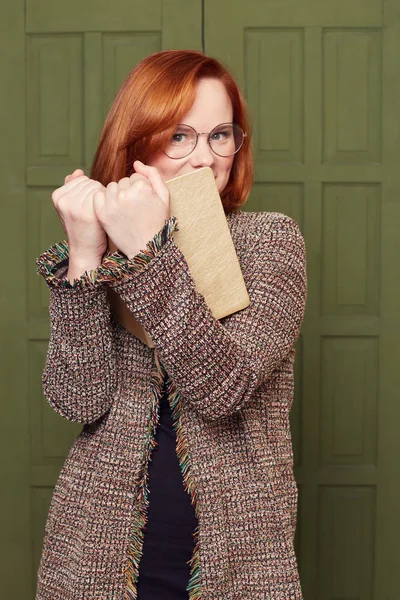 Gorgeous redheaded female book reader hides behind textbook, has foxy face expression, finds out something new, has interesting information, looking at camera from round big spectacles for good vision