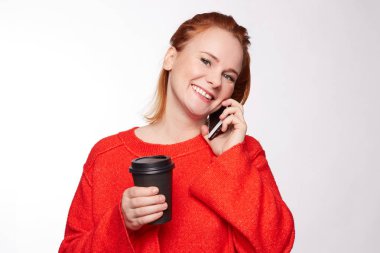 Sevimli gülümseme ile güzel memnun kadın sıcak take away kahve sahiptir, akıllı telefonla arayan, iyi bir ruh hali vardır. Kızıl saçlı güzel beyaz bir kadın kırmızı kazaklı modern cep konuşuyor.
