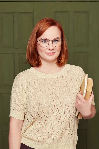 Good looking ginger haired woman with satisfied expression, smiles gently, has intrigued look, thinks about creative plan, studies at college or university, holds books, posing over classroom door.