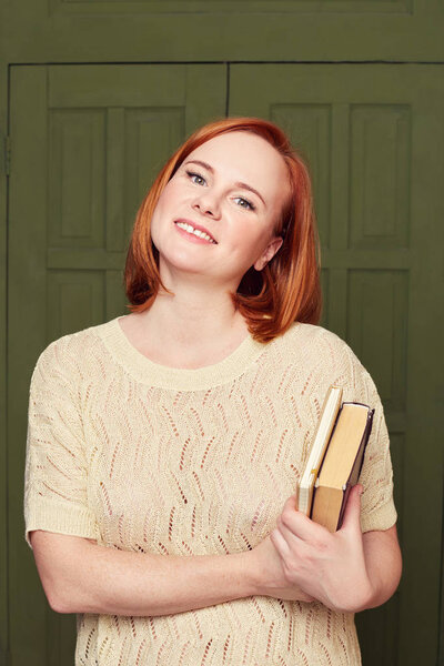 Attractive female student with freckled skin, red hair busy with presentation for seminar, reads book and writes notes, looks joyfully at camera, feeling proud of studying in international university.