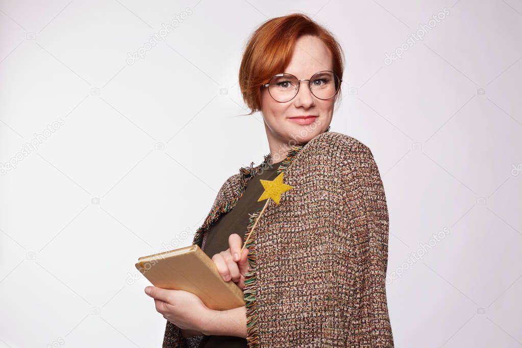 Portrait of successful female writer or journalist, makes notes in diary, holds bookmark near mouth, wears round glasses, has red hair, isolated over white blank studio wall. Youth, education, concept