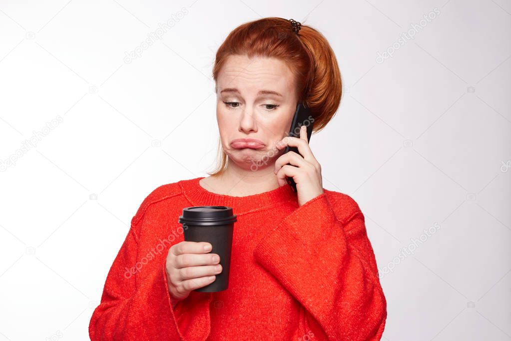 Displeased upset ginger female curves lip, being unhappy to receive bill from banking service on mobile phone, has no money to pay, feels frustrated and disappointed, isolated over white studio wall.