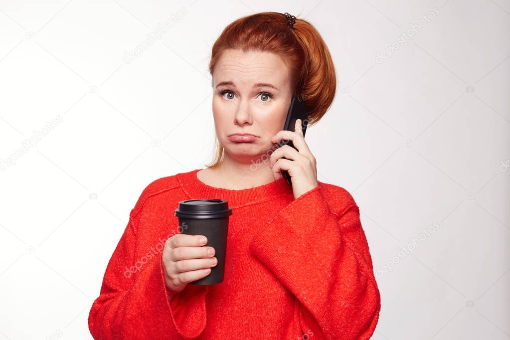 Isolated portrait of beautiful American woman with messy ginger hair, talking on smart phone with sad and disappointed expression, listening to some unexpected news, standing alone over white wall.