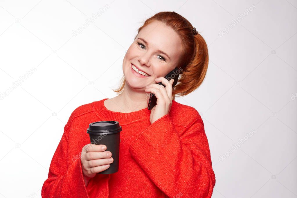 Happy woman dressed casually, chats with friend over smart phone, drinks hot beverage, stands indoors, enjoys good morning. Wife calls husband,asks when he finishes work, has pleasant conversation.