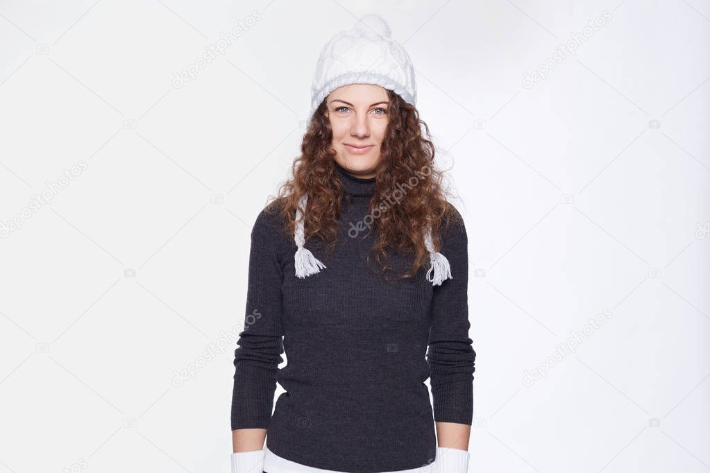 Portrait of attractive blue eyed young Caucasian woman with curly hair, wearing knitted cap with pompon, roll-neck grey sweater, looking confident at camera, being ready for cold times, posing indoors