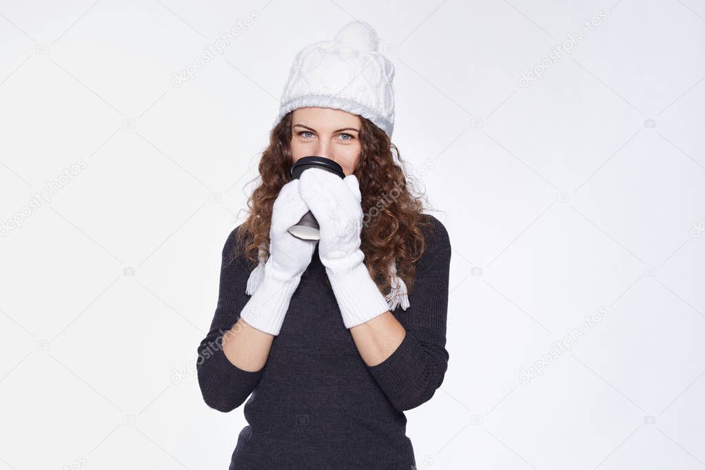 Studio portrait of pretty lady, dressed in trendy white cap with pompon, knitted mittens, grey neck-roll sweater, drinking take away coffee, trying to warm up, looking flirty at camera with blue eyes.