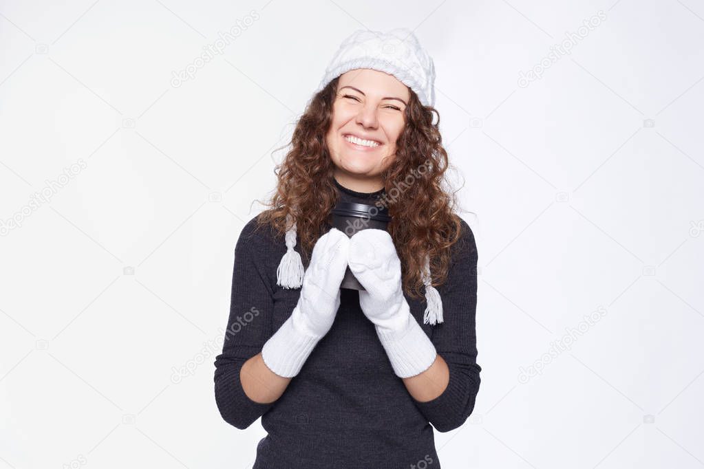 Overjoyed female with perfect smile drinks hot beverage from disposable black paper cup,enjoys spending time in new winter outfit, likes friendly conversation, isolated over white wall with copy space