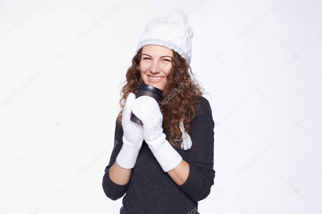 Overjoyed female with perfect smile drinks hot beverage from disposable black paper cup,enjoys spending time in new winter outfit, likes friendly conversation, isolated over white wall with copy space