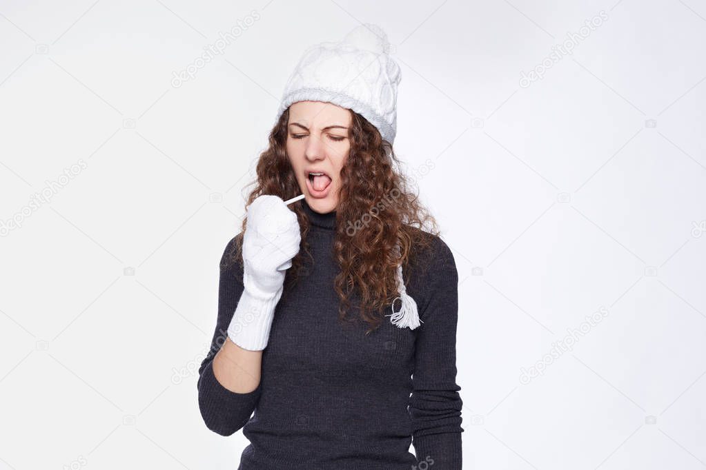 Lovely young curly haired female has pain in throat, suffers from cough attack, keeps hands in mittens, head in hat, mouth opened, has negative facial expression, going to spray disgusting medicine.