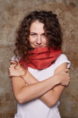Sevimli kıvırcık saçlı kadın titriyor soğuk, yarı uzunluk dikey çekim ateşi var, diş clenches, eller göğüs üzerinde geçti, beyaz tişört ve kaşmir eşarp, beton duvardan izole giyer. 