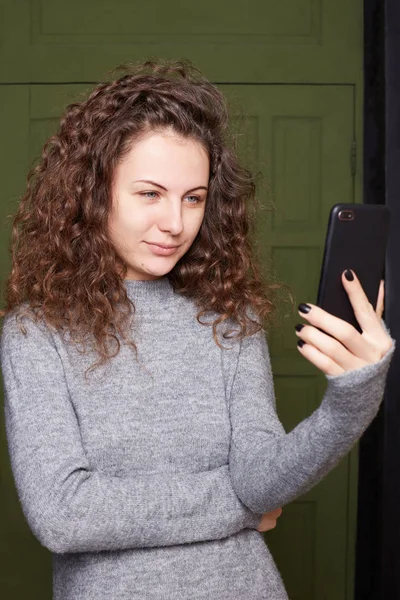 Glad satisfied woman with foxy look poses in front of cell phone camera, makes picture to upload at social networks, takes selfie, dressed in casual roll-neck sweater, isolated over wooden door.