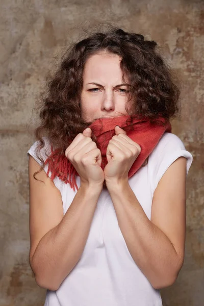 So freezing! Portrait of frustrated displeased Caucasian woman hugs herself, trembles from cold, wears scarf on summer clothes during windy weather, feels discomfort, poses against concrete background