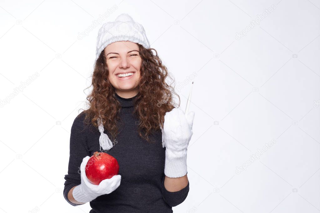 Portrait of beautiful curly haired lady wearing knitted cap and gray roll-neck sweater, winking smiling at camera with happy cheerful look,holding pomegranate and thermometer, being finally healthy.