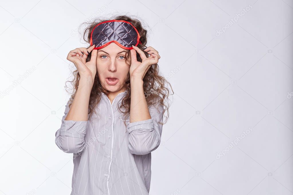 Beautiful female adult grimaces, taking off sleeping mask from face, has sleepy expression, fed up of morning routine, wants to have holidays, dressed in stylish striped pajamas, isolated on white. 
