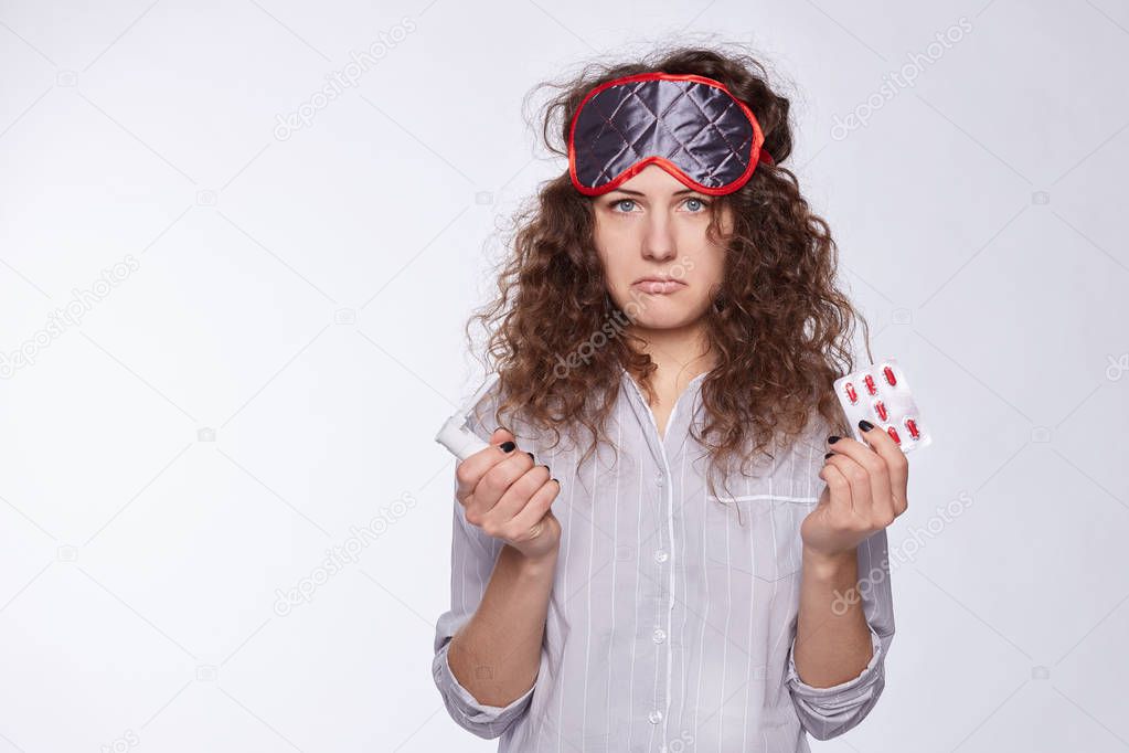 Unhappy stressed young curly haired female with blue eyes holding pills suffering from ache, frowning and looking at camera with tensed and painful expression on pretty face. Health anf body language. 